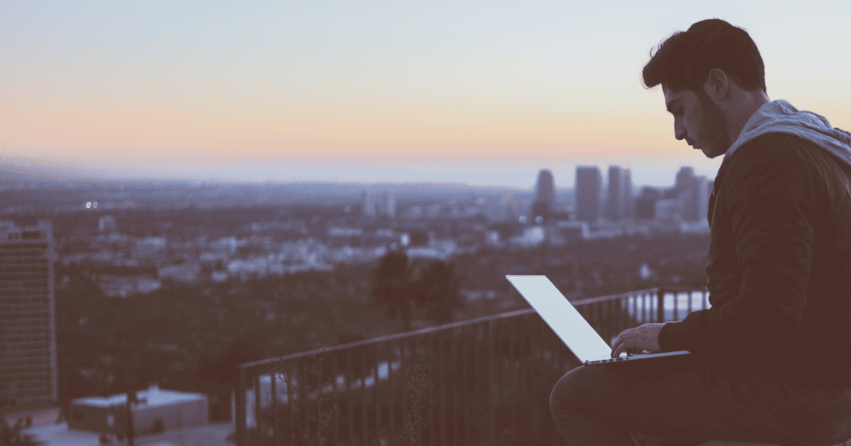 Man sitting on ledge above a city on his computer during sundown.