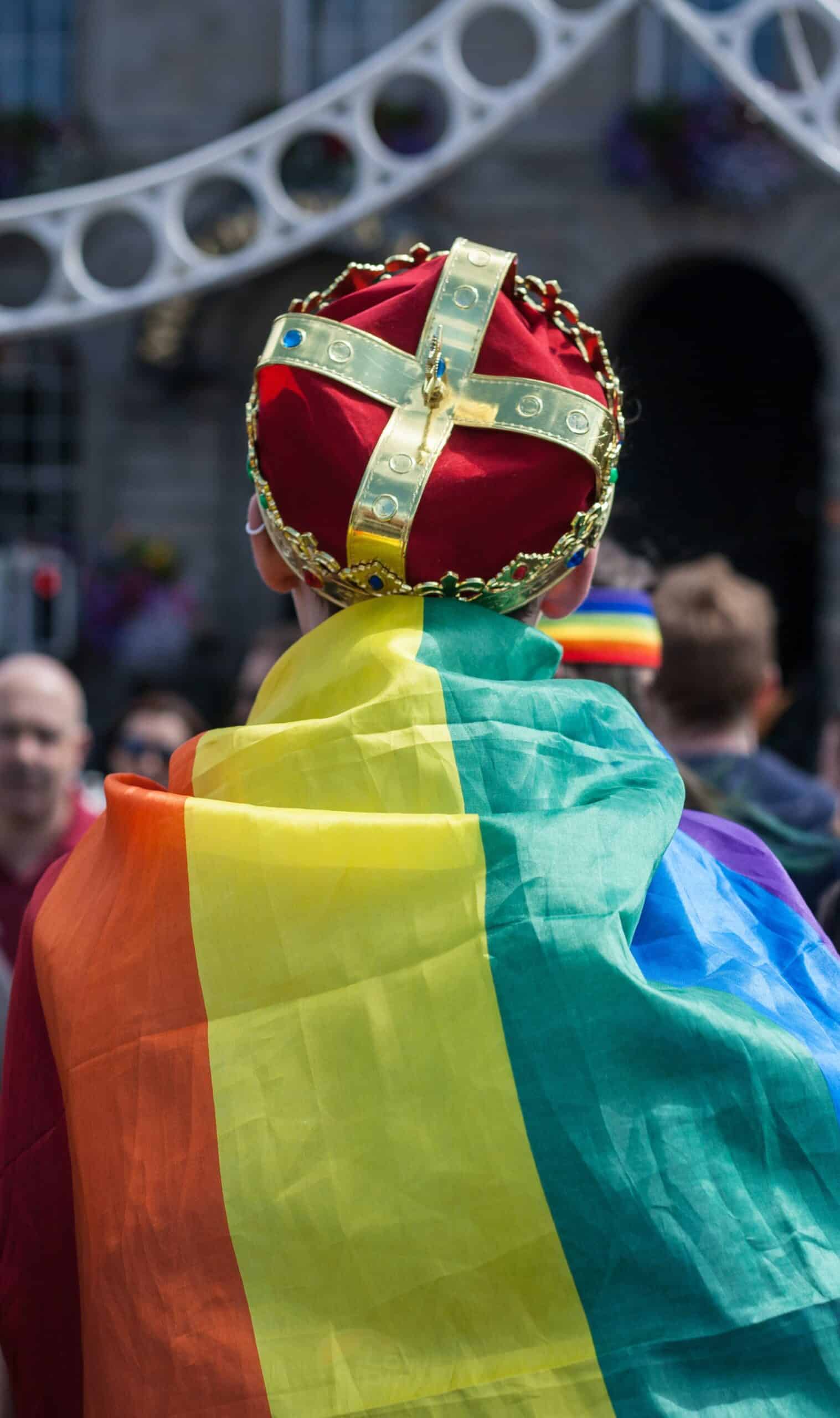 Man wearing a rainbow Pride flag on his back while also wearing a crown. 