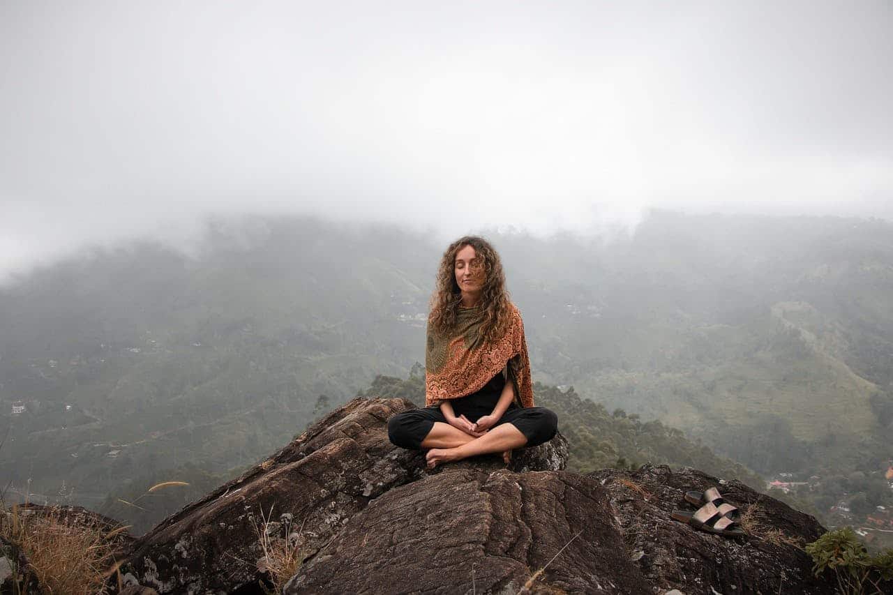 A woman sitting cross-legged on top of a mountain meditating with fog in the background.