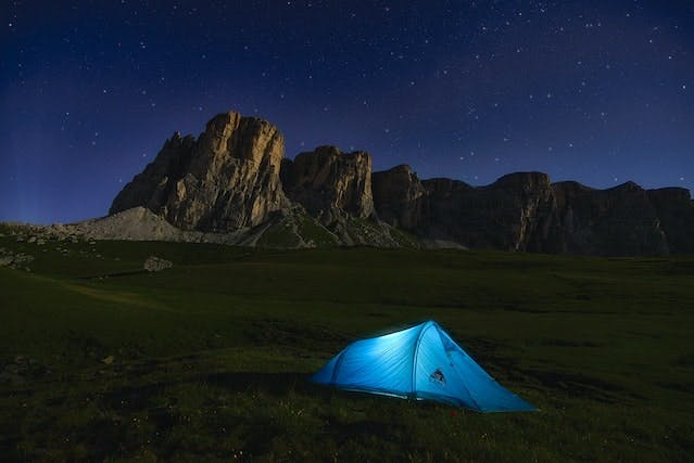 Blue tent with a light on in front of a rock formation and a starry night